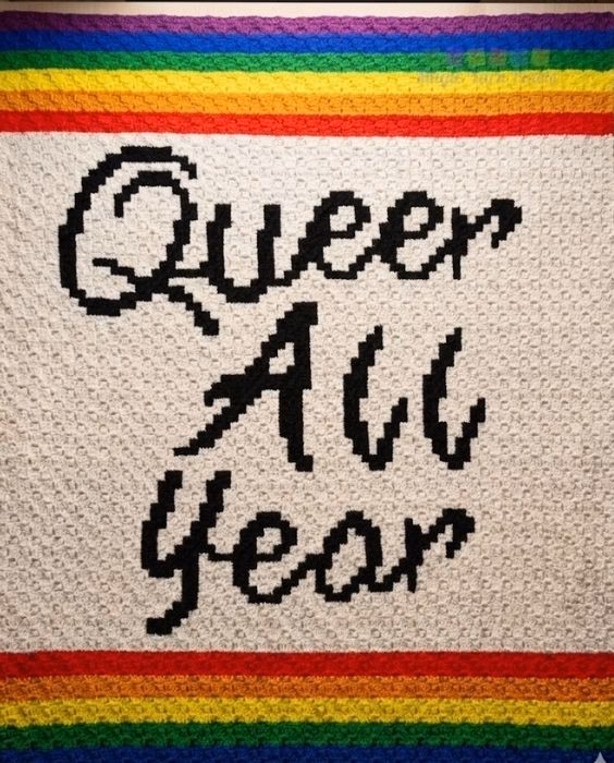 A corner-to-corner crochet blanket laid flat, featuring the text "Queer All Year" in black pixelated letters on a white background, bordered by rainbow stripes at the top and bottom, featured in a crochet roundup from https://magicyarnpixels.com/.