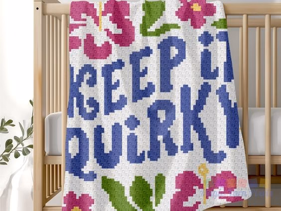 A C2C crochet baby blanket with the text "KEEP IT QUIRKY" in blue, draped over the side of a wooden crib. The blanket features pink hibiscus flowers and green leaf accents on a white background. This image is part of a crochet roundup from https://magicyarnpixels.com/ highlighting c2c crochet designs and graphs.