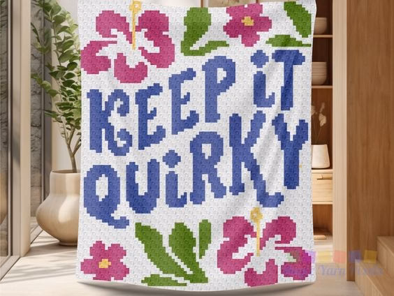 A C2C crochet blanket featuring the text "KEEP IT QUIRKY" in blue, hanging in a bright, modern room with a potted plant and wooden shelving in the background. The blanket is decorated with pink hibiscus flowers and green leaves. This project is part of a crochet roundup from https://magicyarnpixels.com/ showcasing c2c crochet designs and blanket graphs.