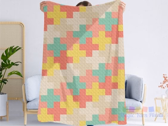 A person holds up a large crochet blanket featuring the Modern Kawaii Crosses pattern. The design consists of repeating, interlocking cross shapes in coral, yellow, mint green, and cream. The blanket is displayed in a bright room with a wooden sofa and a potted plant in the background. A small Magic Yarn Pixels logo is visible in the bottom right corner.