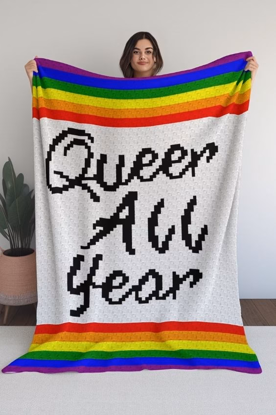 A woman holding up a large crocheted corner-to-corner blanket with the text "Queer All Year" in black pixelated letters on a white background, framed by rainbow stripes at the top and bottom, featured in a crochet roundup from https://magicyarnpixels.com/.