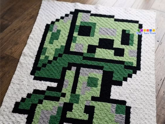 A white corner-to-corner crocheted blanket featuring a pixel art Minecraft Creeper design, shown flat on a wooden floor with a small brand watermark in the top right. Original design by Ariana of MagicYarnPixels.