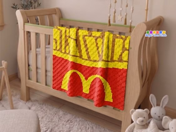 A red corner-to-corner (C2C) crochet baby blanket draped over the side of a wooden crib, showcasing a pixel art design of fast food french fries in a red box with the iconic yellow golden arches logo. Original Design by Ariana of MagicYarnPixels.