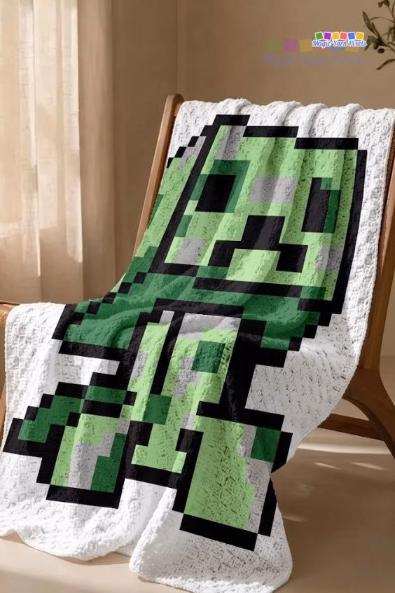 A white corner-to-corner crocheted blanket with a green Minecraft Creeper pixel art design, styled across a wooden accent chair. Original design by Ariana of MagicYarnPixels.