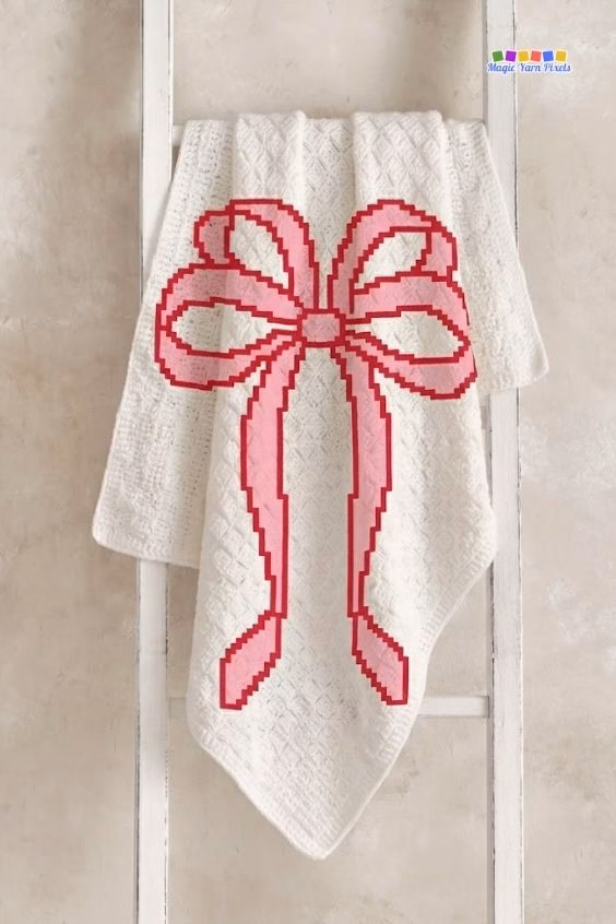 Cream C2C crochet blanket featuring a pink pixel coquette bow design, hanging on a white wooden display ladder. Original design by Ariana of MagicYarnPixels.