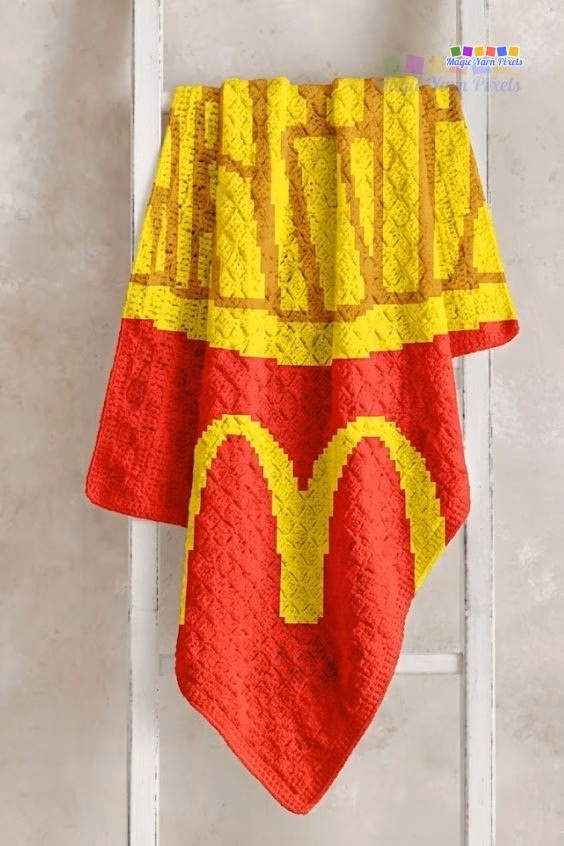 A red corner-to-corner (C2C) crochet blanket hanging over the rungs of a white decorative wooden ladder against a neutral wall, showcasing a pixel art design of fast food french fries in a red box with the iconic yellow golden arches logo. Original Design by Ariana of MagicYarnPixels.