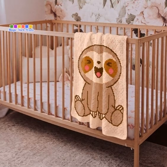 A peach corner-to-corner (C2C) crochet baby blanket with a pixel art baby sloth design draped over a wooden crib. The background is a pink and white floral wallpaper. The Magic Yarn Pixels logo is in the top left corner. Original design by Ariana of MagicYarnPixels.