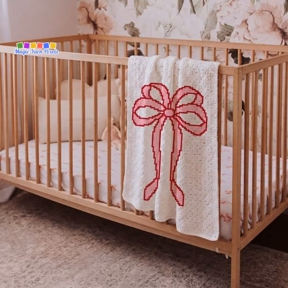 Cream C2C crochet nursery blanket with a large pink pixel coquette bow design draped over a wooden crib rail. Original design by Ariana of MagicYarnPixels.