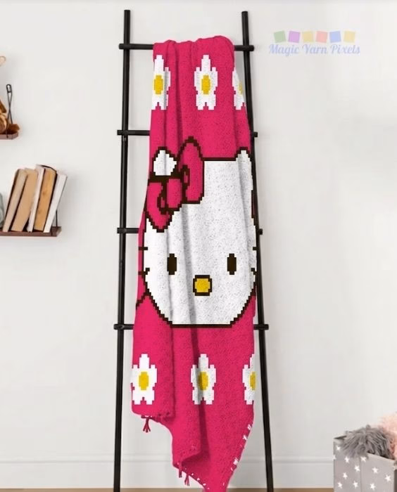 Cute Hello Kitty C2C crochet blanket draped vertically over a black display ladder, showing Hello Kitty's face with a bright pink bow on a hot pink background with white and yellow flowers. Original design created by Ariana Wimsett at MagicYarnPixels.com