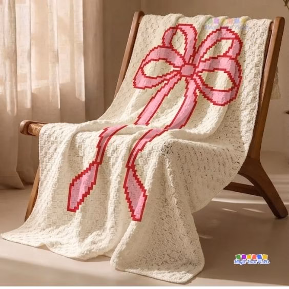 Cream C2C crochet blanket featuring a large pink pixel coquette bow design draped over a wooden chair. Original design by Ariana of MagicYarnPixels.