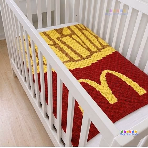 A corner-to-corner (C2C) crochet baby blanket neatly laid inside a white wooden crib, showcasing a pixel art design of fast food french fries in a red container with the iconic yellow golden arches logo. Original Design by Ariana of MagicYarnPixels.