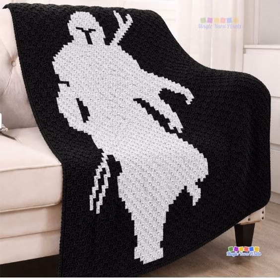 A black and white corner-to-corner crochet blanket featuring a pixel art silhouette of The Mandalorian draped over a couch. Original design by Ariana of MagicYarnPixels.