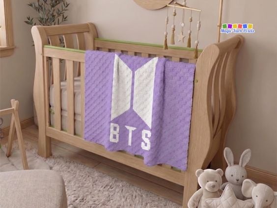 A purple and white corner-to-corner (C2C) crocheted blanket hanging over the side of a light wooden crib in a nursery room. The blanket features a prominent white BTS geometric logo with the block letters "BTS" stitched below it. Stuffed animals sit on the floor beside the crib. Original design by Ariana of MagicYarnPixels.