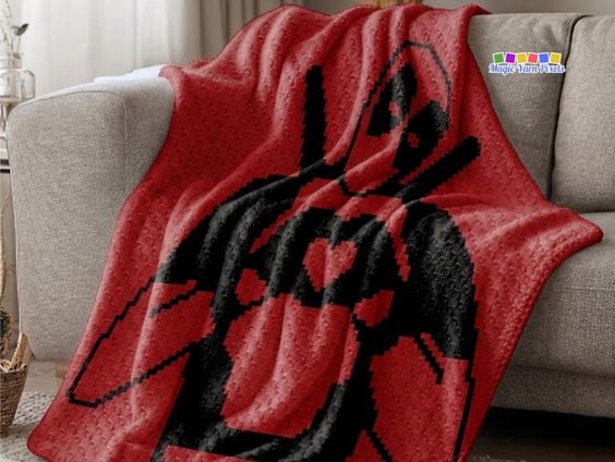 A red corner-to-corner (C2C) crochet blanket draped over a gray couch, displaying a large black silhouette design of Deadpool making a heart shape with his hands. Original design by Ariana of MagicYarnPixels