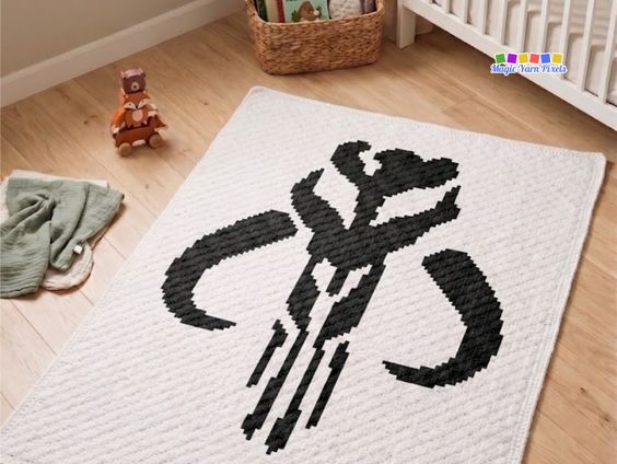 A corner view of a bright nursery featuring a cream-colored crochet blanket spread flat on a light wood floor. The blanket showcases a bold, black Mandalorian crest Mythosaur skull pixel design. Nearby on the floor is a small stack of wooden fox toys and a folded green muslin cloth, with a wicker basket sitting next to a white wooden crib in the background. A "Magic Yarn Pixels" logo is visible in the top right corner. Original design by Ariana of MagicYarnPixels.
