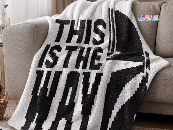 A close-up of a white and black crochet blanket draped over a grey couch. It features a large pixel art design of a Mandalorian helmet and the words "THIS IS THE WAY". Original design by Ariana of MagicYarnPixels.
