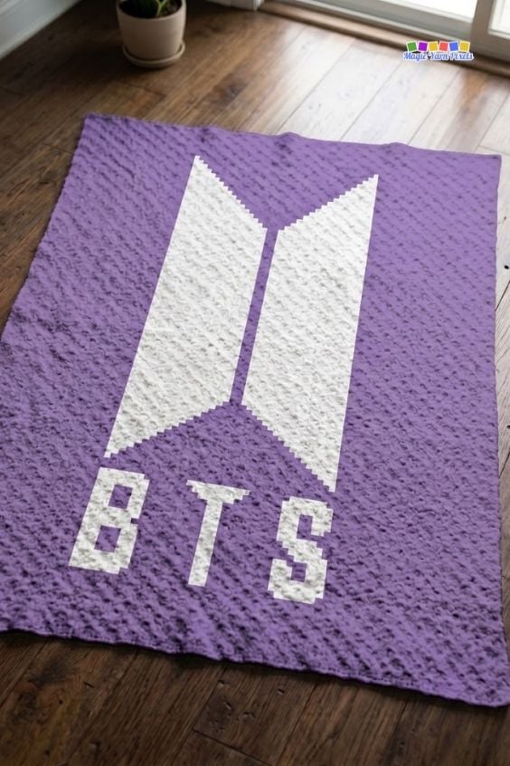 A purple and white corner-to-corner (C2C) crocheted blanket laid flat on a dark wooden floor. The blanket showcases the white geometric BTS logo centered above the pixelated block letters "BTS" against a solid purple textured background. Original design by Ariana of MagicYarnPixels.