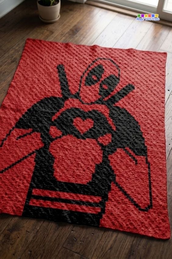 A red corner-to-corner (C2C) crochet blanket laid out flat on a polished hardwood floor, showcasing a black silhouette design of Deadpool making a heart shape with his hands. Original design by Ariana of MagicYarnPixels.