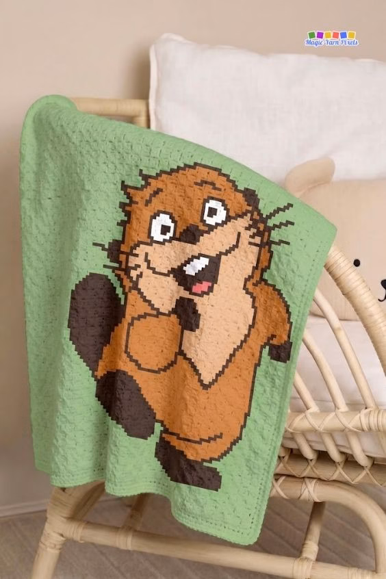 A green crochet blanket hanging over the side of a light-colored wicker crib. The blanket features a large, pixel-style cartoon beaver smiling and waving. Original design by Ariana of MagicYarnPixels.