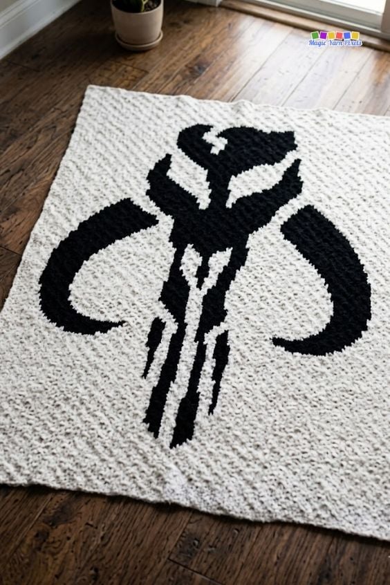 A high-angle shot focusing on a cream corner-to-corner crochet blanket laid out on a dark hardwood floor. The blanket features a prominent black pixelated Mandalorian crest Mythosaur skull design. A potted plant sits in the upper left corner on the floor, and a small "Magic Yarn Pixels" logo is placed in the top right. Original design by Ariana of MagicYarnPixels.