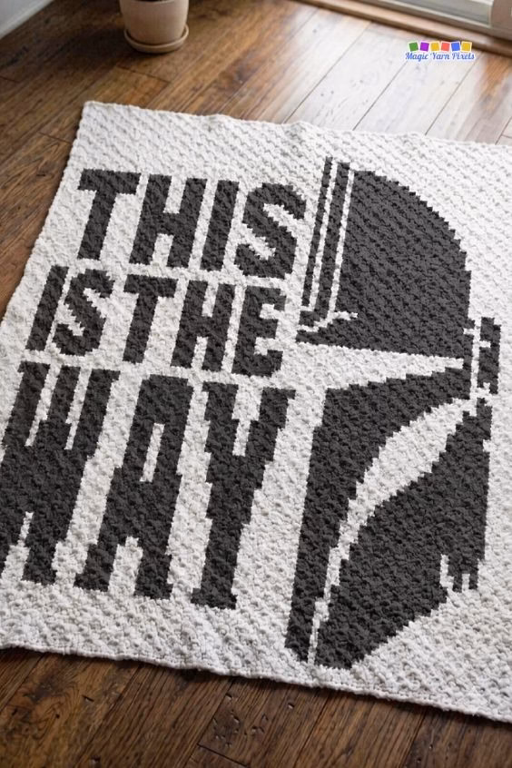 A white crochet blanket spread out flat on a dark wooden floor. It features a large black pixel art design of a Mandalorian helmet and the words "THIS IS THE WAY". Original design by Ariana of MagicYarnPixels.