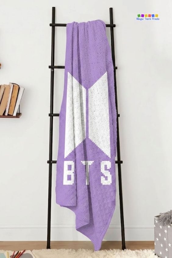 A purple and white corner-to-corner (C2C) crocheted blanket hanging vertically from a black decorative ladder against a plain white wall. The folds of the blanket show the large white geometric BTS logo and the block text "BTS" underneath it. Original design by Ariana of MagicYarnPixels.