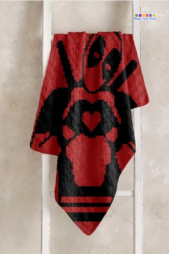 A red corner-to-corner (C2C) crochet blanket hanging vertically from a white wooden display ladder against a textured wall, highlighting the black silhouette design of Deadpool making a heart shape with his hands. Original design by Ariana of MagicYarnPixels.