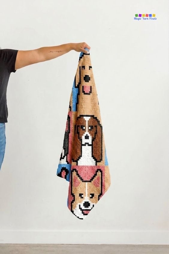 An arm extending from the left holds up a folded, vertical strip of a colorful crochet blanket against a plain white wall. The section on display shows three pixel-art dog faces stacked vertically. A small "Magic Yarn Pixels" logo is visible in the top right corner. Original design by Ariana of MagicYarnPixels.