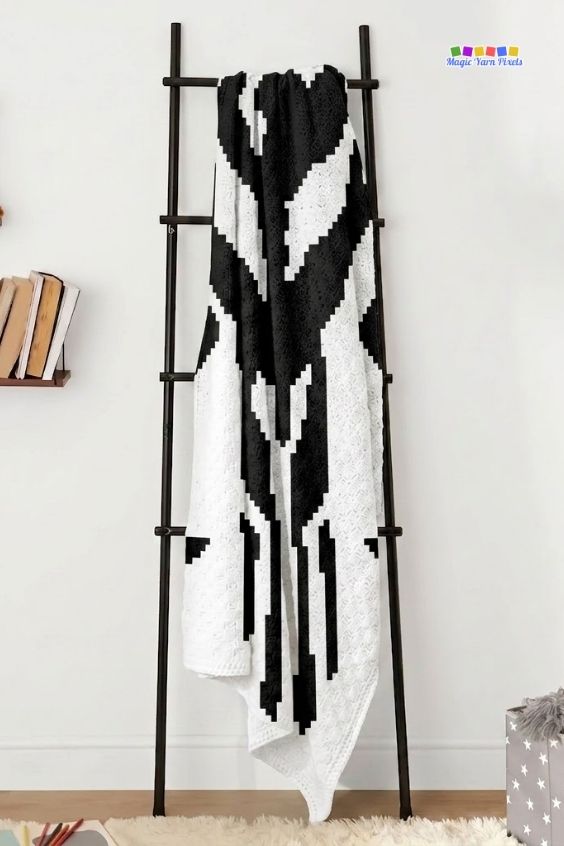 A cream and black crochet blanket draped vertically over a dark brown decorative blanket ladder against a plain white wall. The section of the blanket on display highlights the geometric, pixelated lines of the black Mandalorian crest Mythosaur skull design. A small wall shelf with books is visible on the left, and a "Magic Yarn Pixels" logo is in the top right corner. Original design by Ariana of MagicYarnPixels.
