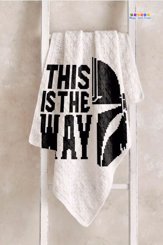 A white crochet blanket hanging on a light wood ladder against a textured wall. It features a large black pixel art design of a Mandalorian helmet and the words "THIS IS THE WAY". Original design by Ariana of MagicYarnPixels.