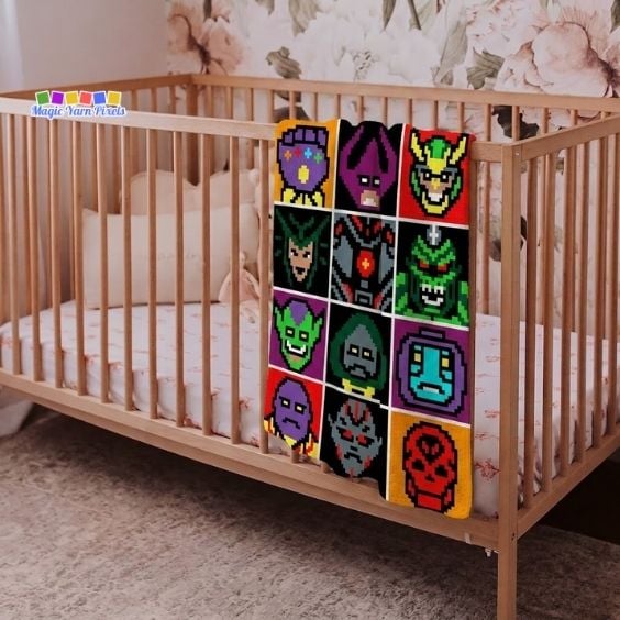 A colorful corner-to-corner (C2C) crochet blanket hanging over the side of a wooden crib in a nursery, displaying a grid pattern of twelve distinct pixel art character blocks of Avengers villains. Original design by Ariana of MagicYarnPixels.
