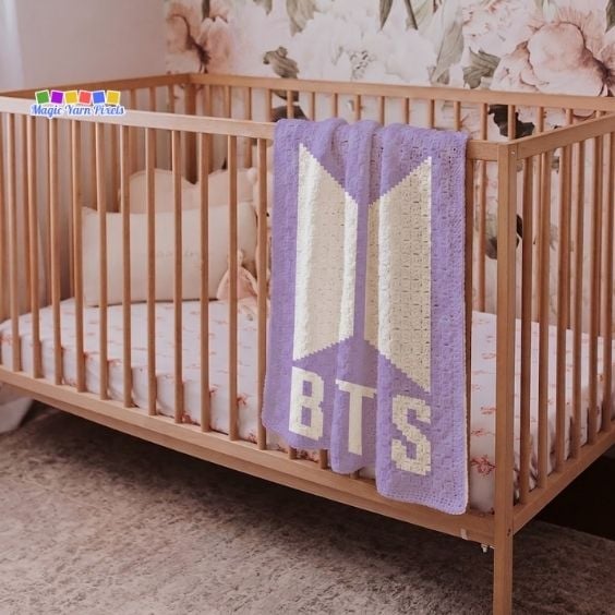 A purple and white corner-to-corner (C2C) crocheted blanket draped over the railing of a light wooden crib. The blanket features the distinct white pixelated BTS logo alongside the text "BTS" across the lower portion. The background shows a nursery room with floral patterned wallpaper. Original design by Ariana of MagicYarnPixels.