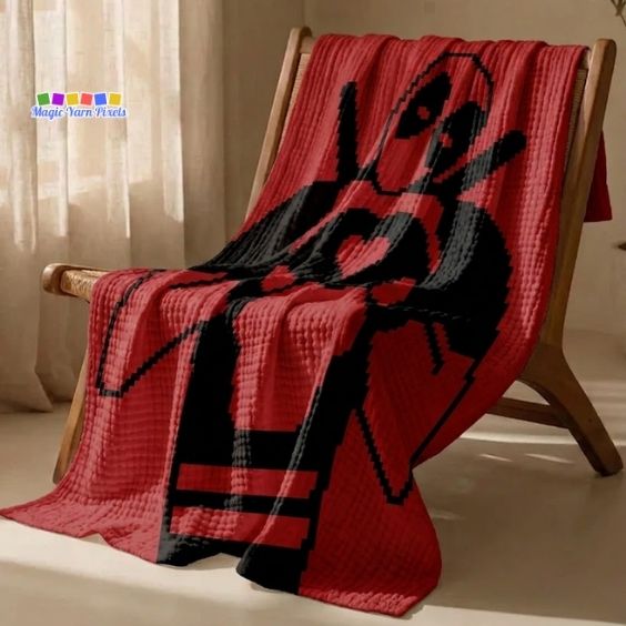 A red corner-to-corner (C2C) crochet blanket draped comfortably over a wooden armchair, showing the black silhouette design of Deadpool making a heart shape with his hands. Original design by Ariana of MagicYarnPixels.
