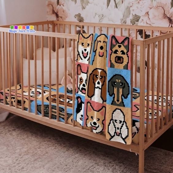 A side-angle view of a wooden crib in a nursery with a colorful crochet blanket draped over the side rail. The blanket features a grid of nine pixelated dog breed faces. The crib is lined with a mattress cover featuring a matching dog pattern, and a muted floral wallpaper decorates the wall behind it. A "Magic Yarn Pixels" logo is in the top left corner. Original design by Ariana of MagicYarnPixels.