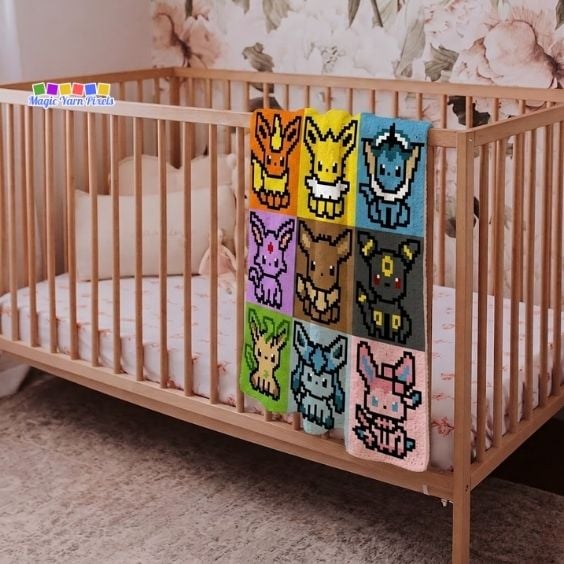 A C2C crochet blanket featuring nine pixelated Eevee evolutions draped over the side rail of a wooden crib. The nursery background features soft floral wallpaper, a plush white pillow inside the crib, and a soft beige carpet on the floor. The Magic Yarn Pixels logo is located on the top left. Original design by Ariana of MagicYarnPixels.