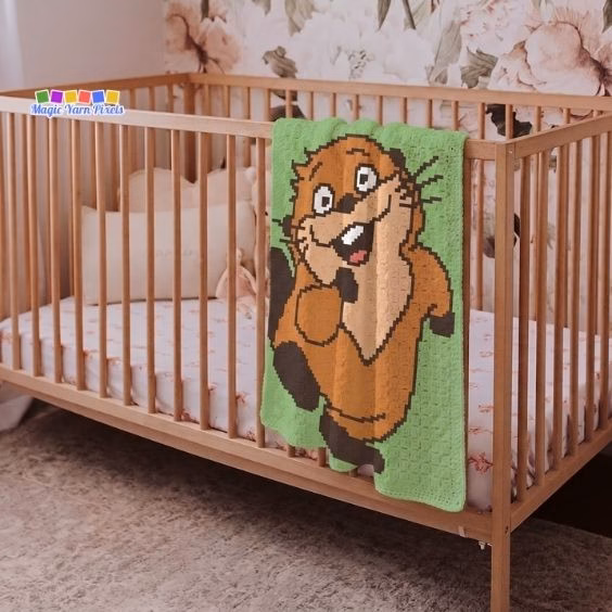 A green crochet blanket hanging over the side of a wooden baby crib. The blanket features a large, pixel-style cartoon beaver smiling and waving. Original design by Ariana of MagicYarnPixels.