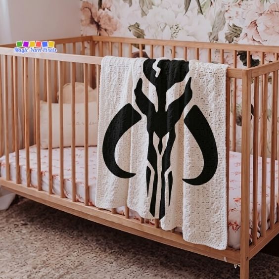 A close-up side view of a cream-colored textured crochet blanket draped over the side of a wooden crib in a nursery. The blanket features a large black Mandalorian crest Mythosaur skull design. The nursery wall behind the crib features a muted floral wallpaper pattern. A "Magic Yarn Pixels" logo is positioned in the top left corner. Original design by Ariana of MagicYarnPixels.