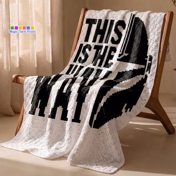A white crochet blanket draped over a wooden lounge chair in a bright room. It features a large black pixel art design of a Mandalorian helmet and the words "THIS IS THE WAY". Original design by Ariana of MagicYarnPixels.
