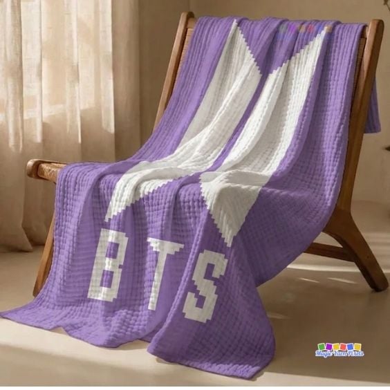 A purple and white corner-to-corner (C2C) crocheted blanket draped over a wooden chair in a bright room. The blanket features the geometric BTS logo in white against a solid purple background, with the block letters "BTS" stitched in white across the bottom. Original design by Ariana of MagicYarnPixels.