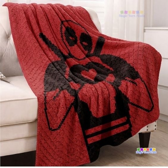 A red corner-to-corner (C2C) crochet blanket draped over the side of a white armchair, prominently displaying a large black silhouette design of Deadpool forming a heart shape with his hands. Original design by Ariana of MagicYarnPixels.