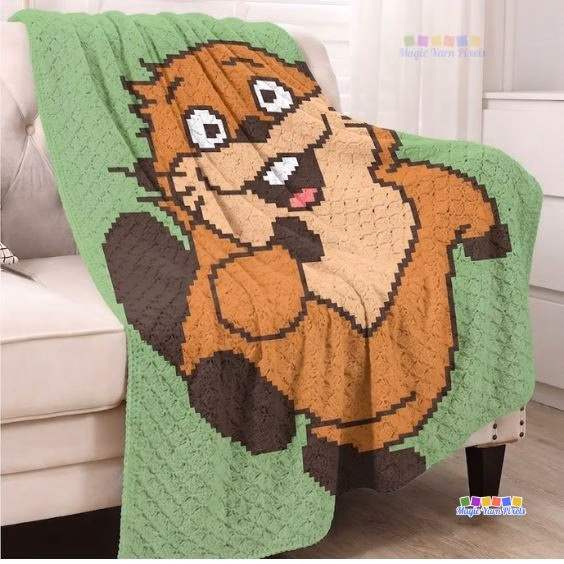 A green crochet blanket draped over a white armchair. The blanket features a large, pixel-style cartoon beaver smiling and waving. Original design by Ariana of MagicYarnPixels.
