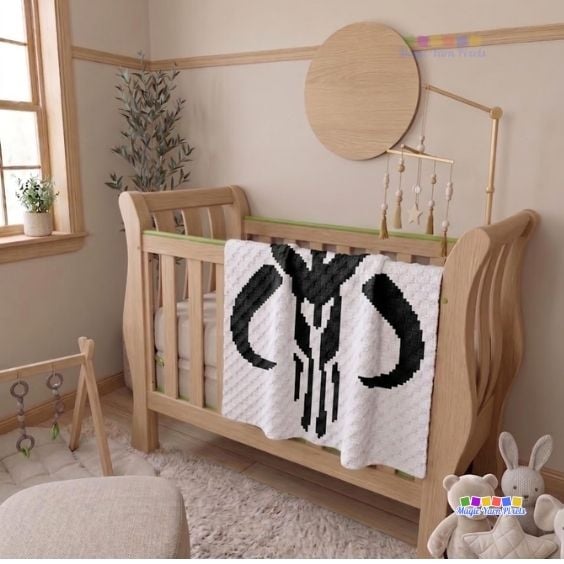 A warm, neutral-toned nursery showing a cream corner-to-corner crochet blanket draped neatly over the railing of a light wood crib. The blanket proudly displays the black Mandalorian crest Mythosaur skull pattern. Stuffed animal toys sit on the floor next to the crib, and a wooden mobile hangs above it. A "Magic Yarn Pixels" logo is in the bottom right corner. Original design by Ariana of MagicYarnPixels.