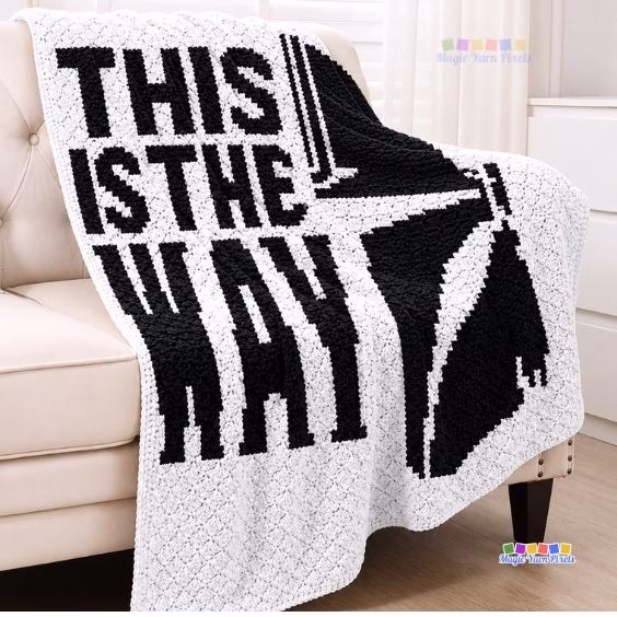 A white and black crochet Star Wars blanket on a chair that says "THIS IS THE WAY" next to a Mandalorian helmet. Original design by Ariana of MagicYarnPixels.