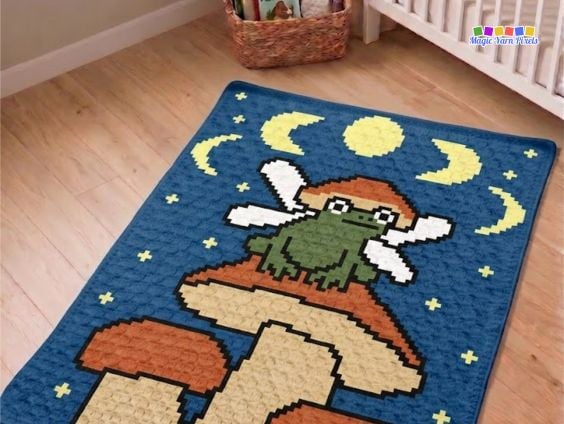 Fairy Frog C2C crochet blanket displayed on a wooden floor, featuring a fairy frog sitting on mushrooms beneath the moon and stars. Original design by Ariana of MagicYarnPixels.