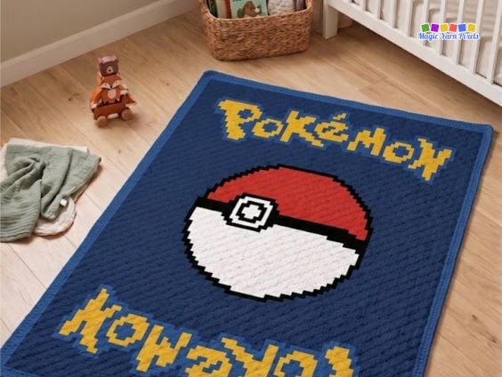 C2C crochet blanket featuring a Pokémon card-inspired design with a centered Poké Ball, yellow pixel-style lettering, and a blue border, displayed on a nursery floor. Original design by Ariana of MagicYarnPixels.