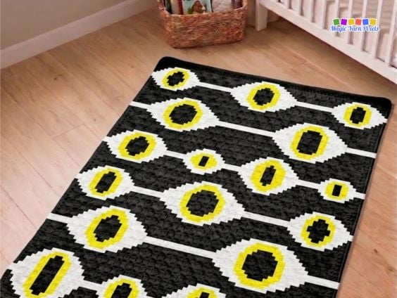 Witchcraft Evil Eye C2C crochet blanket displayed on a wooden floor, showing a repeating evil eye pattern in black, white, and yellow. Original design by Ariana of MagicYarnPixels.