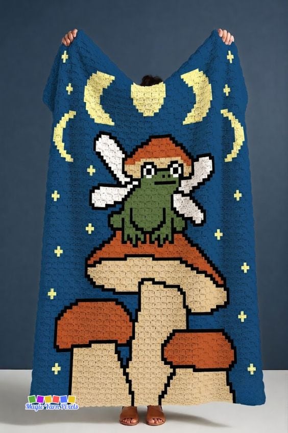 Person holding a Fairy Frog C2C crochet blanket featuring a winged frog on mushrooms under a moonlit sky. Original design by Ariana of MagicYarnPixels.