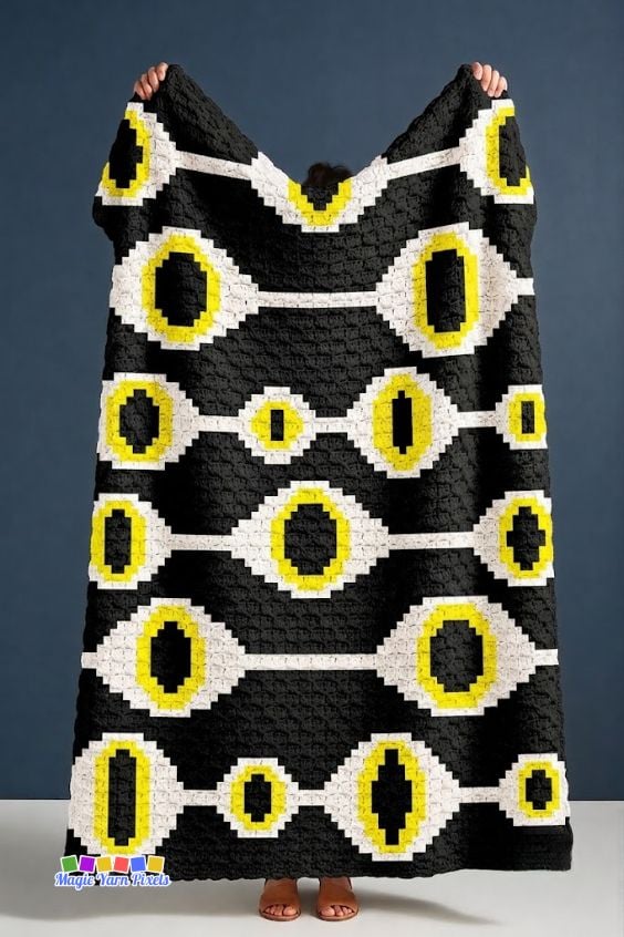Person holding a finished Witchcraft Evil Eye C2C crochet blanket with bold repeating evil eye motifs in black, white, and yellow. Original design by Ariana of MagicYarnPixels.