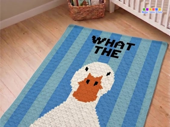 Finished C2C crochet blanket spread across a nursery floor near a crib. The design shows a white duck with an orange bill against blue vertical stripes with bold black text reading "WHAT THE" at the top. Original design by Ariana of MagicYarnPixels.