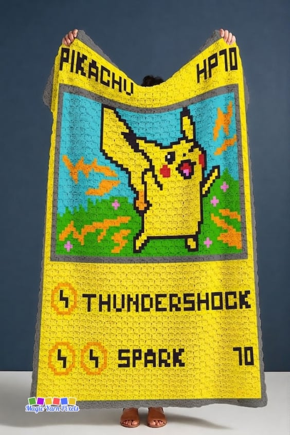 A person holds a finished Pikachu Pok&eacute;mon Card C2C crochet blanket against a dark blue backdrop. The blanket recreates a Pok&eacute;mon trading card with Pikachu, "PIKACHU," "HP70," lightning graphics, and the attacks "THUNDERSHOCK" and "SPARK," finished with a gray border. Original design by Ariana of MagicYarnPixels.